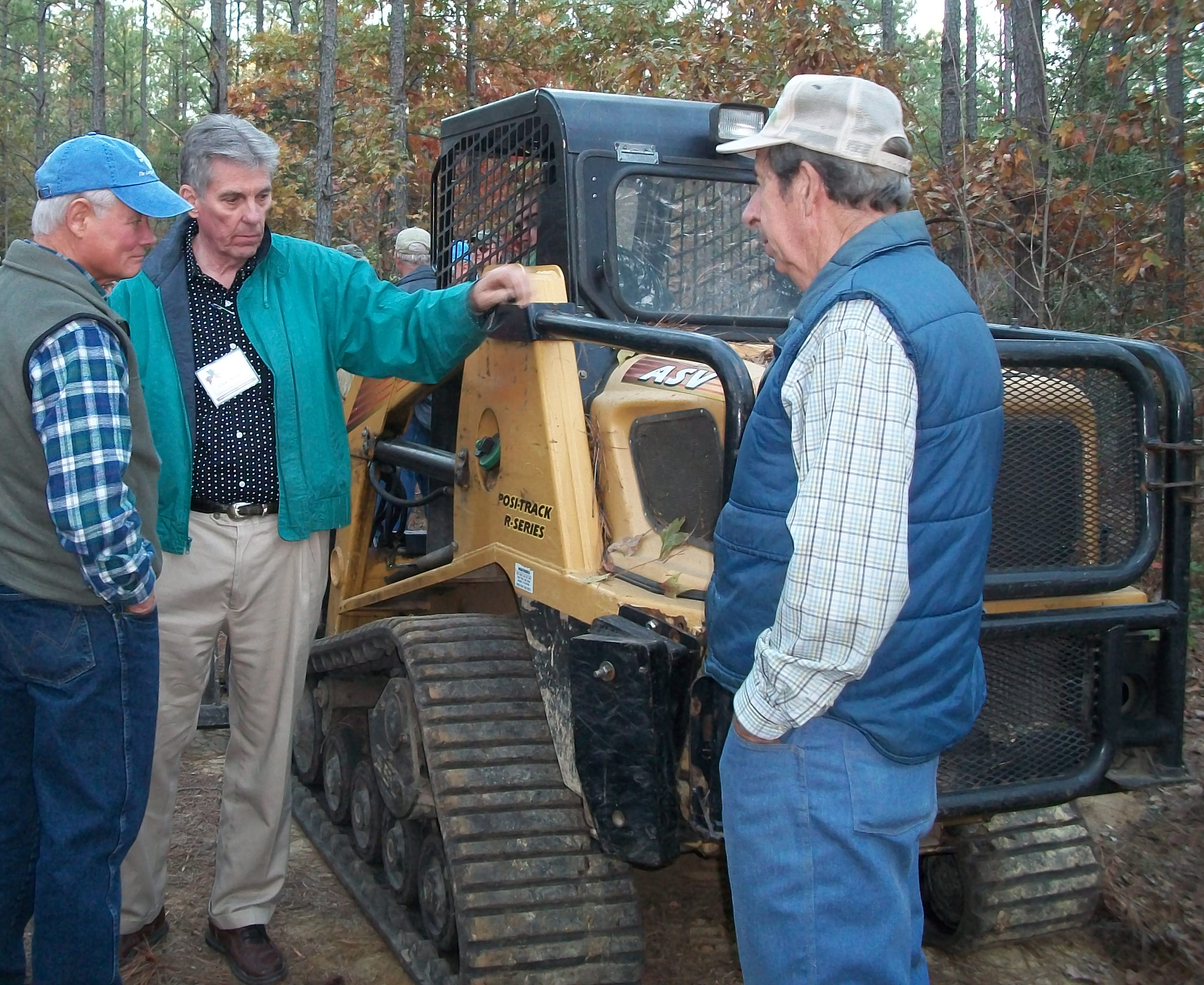 Three men stand looking at a small-scale skidder in a forest.