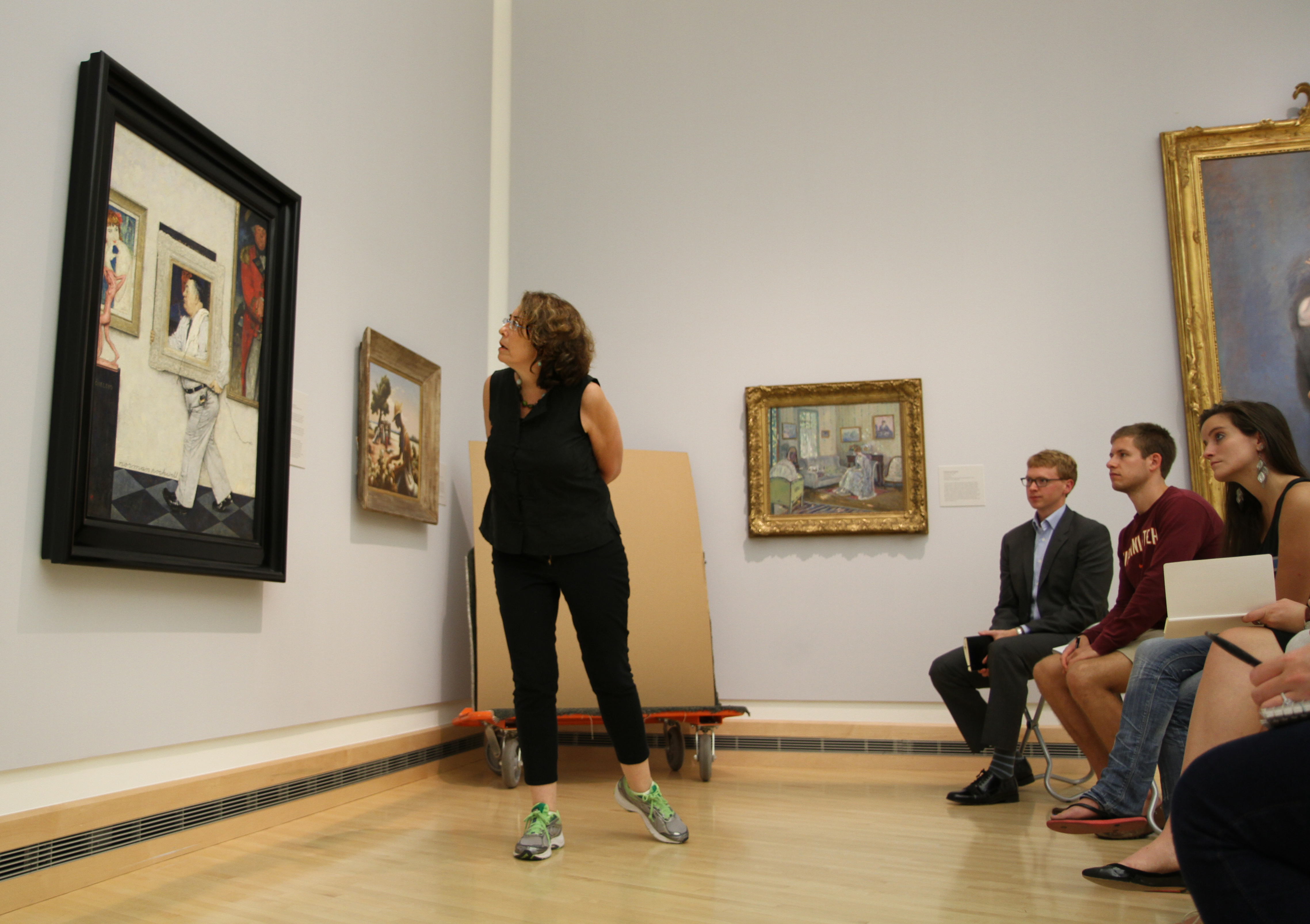 A woman examines a painting on the wall of a museum as seated art history students observe.