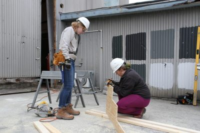 Two female college students in hard hats mark wood to cut. 