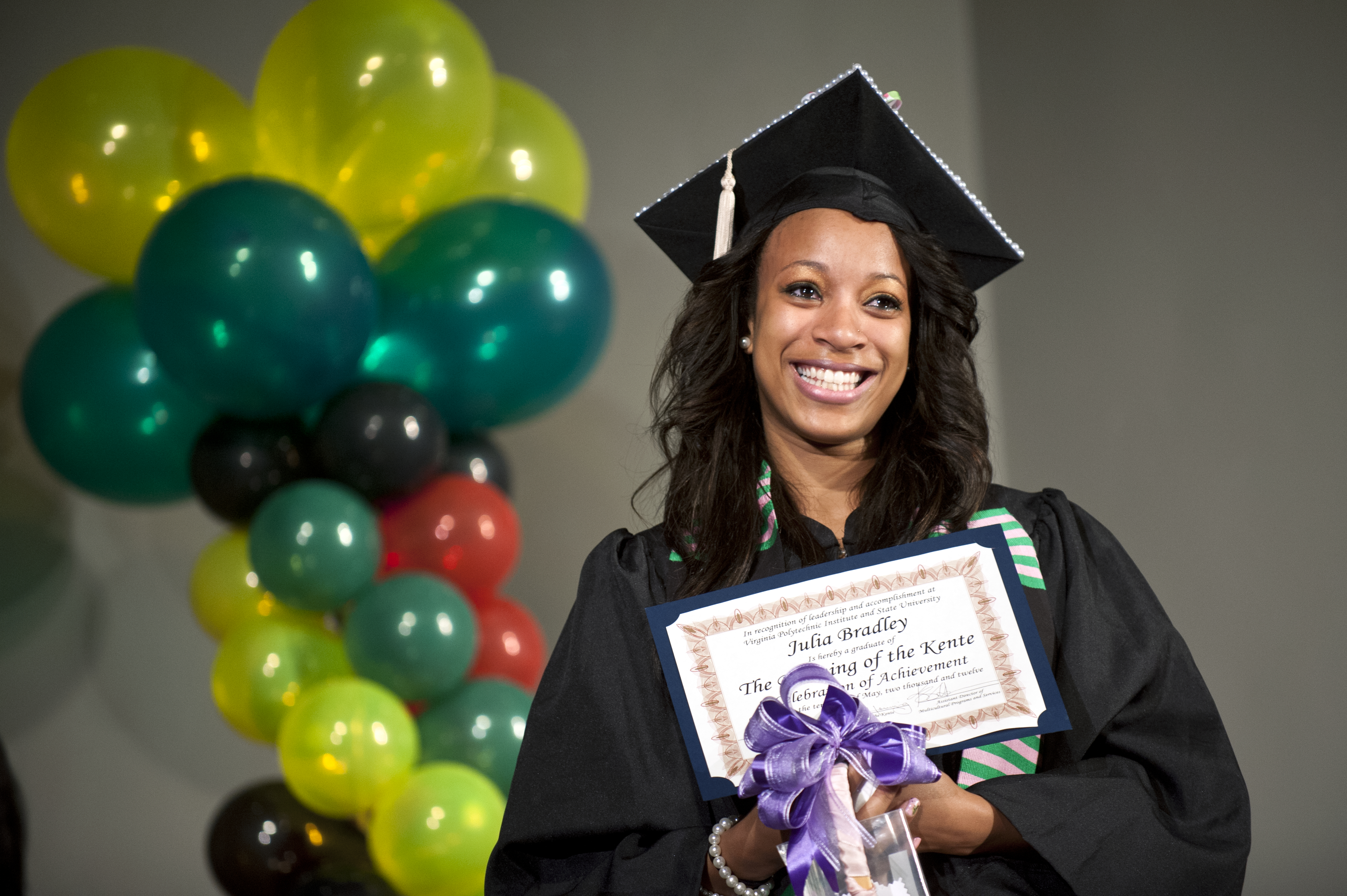 A student dress in commencement regalia smiles with tears in her eyes, as she holds a certificate at the Donning of the Kente ceremony.