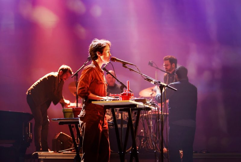Musician Caroline Shaw (front) sings into a microphone with her eyes closed. She has short hair and wears a dark orange jumpsuit. Behind her, the four male members of Sō Percussion play various percussion instruments. A soft, out-of-focus purple hue is projected behind them.