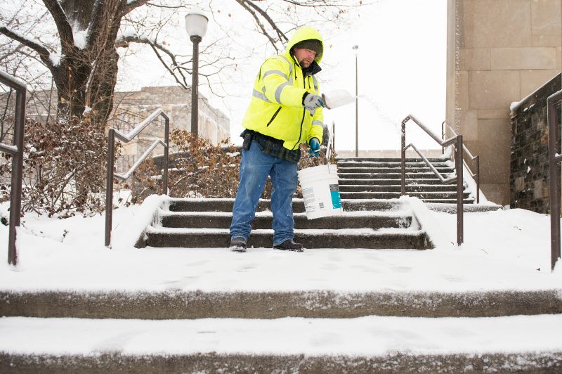Man salts snow-covered cement stairs with a large scoop.